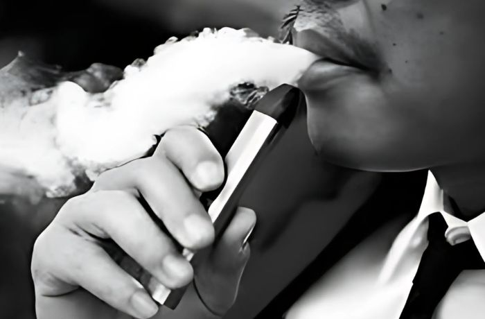 Black and white close-up of a person in formal attire vaping, with smoke being exhaled from their mouth.