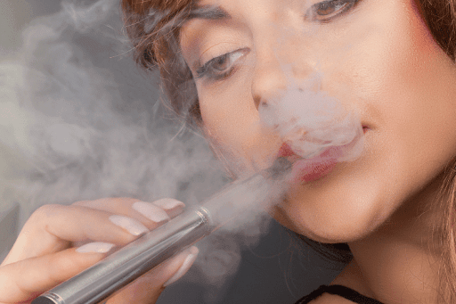 Close-up of a woman exhaling vapor while holding a silver vape pen, surrounded by smoke.