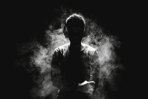 Silhouetted man surrounded by thick smoke, holding a vape device in a dark, moody setting.