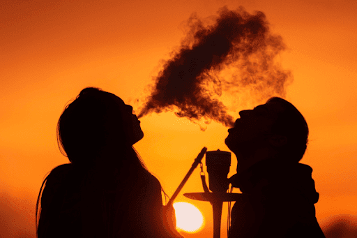 Silhouettes of a man and woman exhaling hookah smoke together against a vibrant orange sunset.