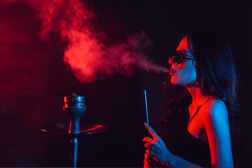 A woman wearing sunglasses smokes hookah in a dimly lit setting with red and blue lighting, creating a dramatic and moody atmosphere.