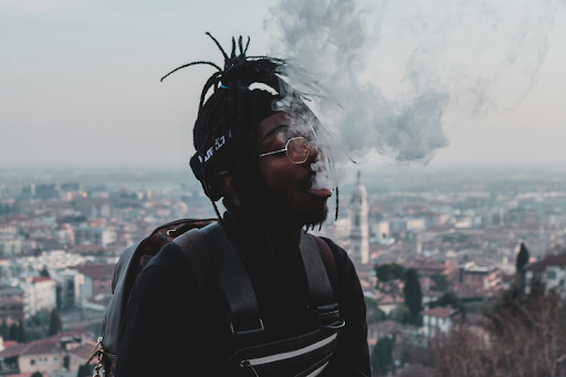 Man with dreadlocks and glasses exhaling vape smoke with a cityscape in the background.