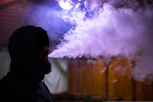 Man in a hoodie exhaling a large cloud of vape smoke indoors, with purple lighting effects.