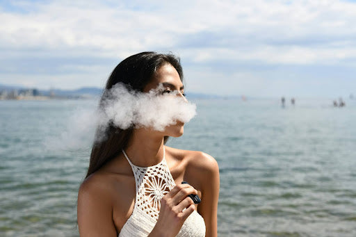 Woman in a white top vaping by the ocean, exhaling a cloud of smoke with the sea and sky in the background.